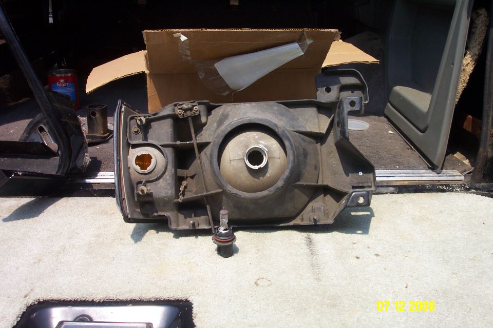 Headlight assembly removed from a vehicle, sitting on a carpeted surface with a cardboard box in the background