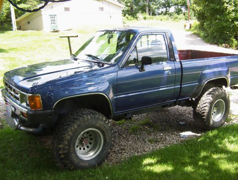 1986 Toyota Hilux - This blue pickup truck is parked on a gravel driveway next to a grassy area with a building in the background.