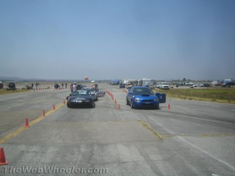 1985 Ford Mustang - Two cars, a blue Subaru WRX and a black Mazda Miata, are lined up on a race track with cones marking the course.