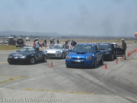 1985 Ford Mustang - Cars are staged on a race track, with cones marking the course and spectators in the background.