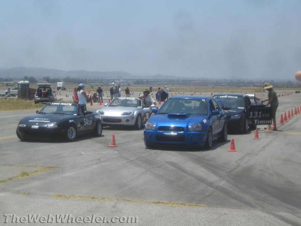 Several cars lined up on a race track, including a blue Subaru WRX, a black Mazda Miata, and a silver Mazda Miata. People are standing around the vehicles