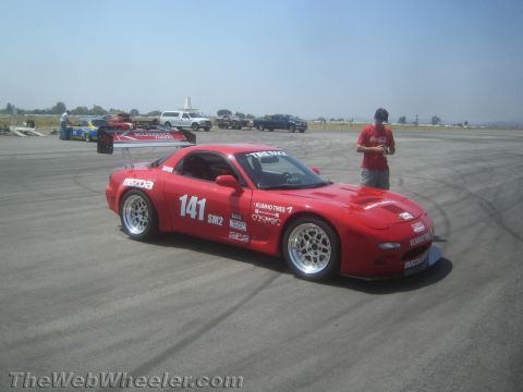 1985 Ford Mustang - A red Mazda RX-7 race car is parked on an asphalt track with other vehicles in the background.