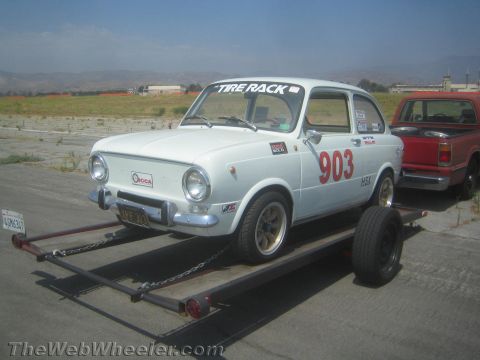 1985 Ford Mustang - A white Fiat 850 Coupe, numbered 903 and bearing 'Tire Rack' branding, is loaded onto a trailer, with a red pickup truck visible behind it.