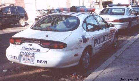 1998 Ford Taurus - A police car is parked on the street next to a sidewalk, with another police car behind it.