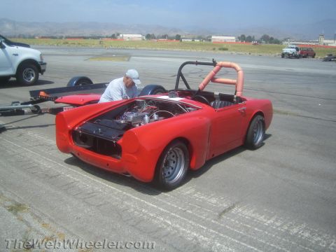 1985 Ford Mustang - A man works on the engine of a red convertible sports car parked on an asphalt surface, possibly an airfield.