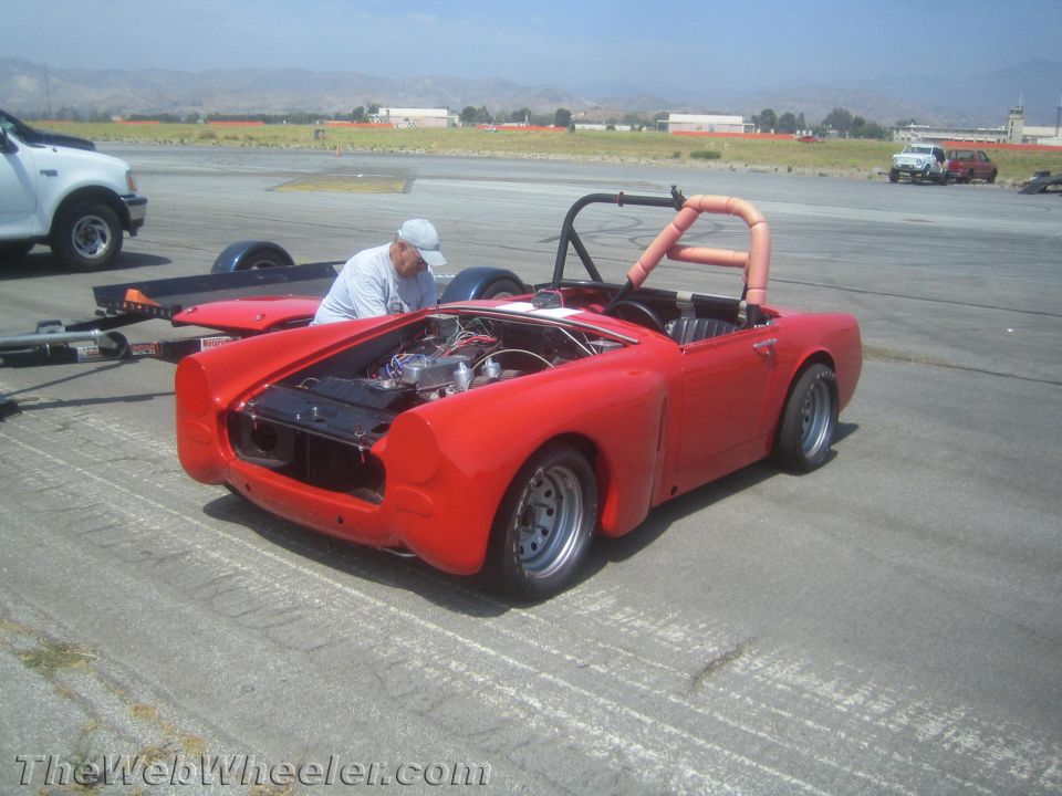 Red convertible sports car, hood open, engine visible. Guy in hat looking at engine. Trailer behind it. White truck nearby