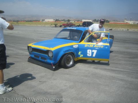 1985 Ford Mustang - A blue and yellow race car, modified for auto racing, is parked on asphalt with a man standing by the open door.