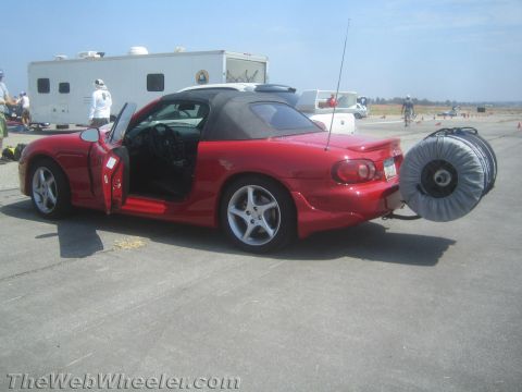 1985 Ford Mustang - Red convertible sports car with a large reel attached to the rear hitch, parked outdoors on a sunny day.