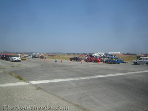 1985 Ford Mustang - Multiple cars are gathered on a race track under a clear blue sky.