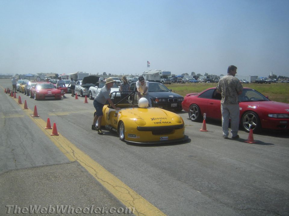 Yellow convertible sports car with Hoosier Tire Rack decals, being pushed by a person. Other cars and people are in the background at a race event