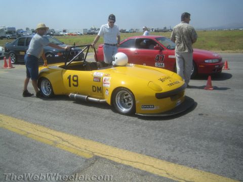 1985 Ford Mustang - A yellow sports car with racing decals sits on the asphalt, possibly at a track event, with spectators around.