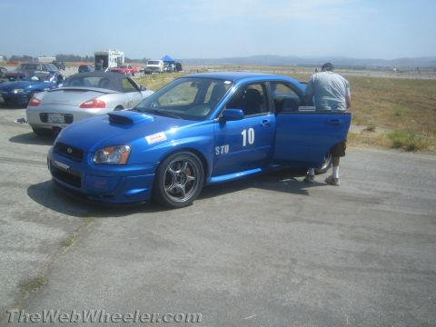 1985 Ford Mustang - A blue Subaru Impreza WRX STI is parked on a racetrack with its driver's door open.