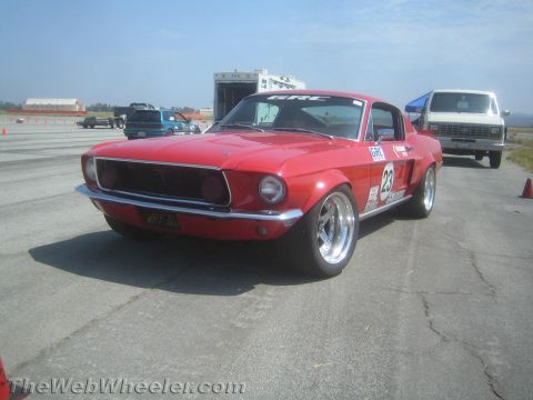 1985 Ford Mustang - A vintage red Ford Mustang, likely from the late 1960s, is prepared for a track event with racing numbers displayed.