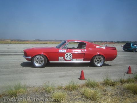 1985 Ford Mustang - A red 1967-1968 Ford Mustang fastback is seen on a race track with other vehicles in the background.