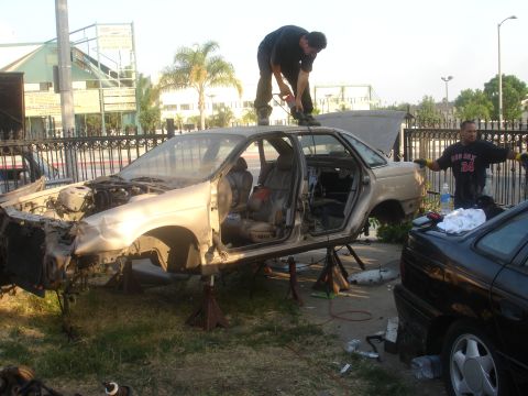 1991 Ford Taurus - A stripped-down car sits on jack stands as two men work on it, with another vehicle nearby.