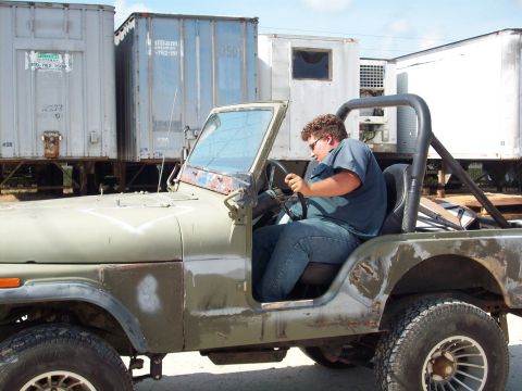 1978 Jeep CJ5 - A man sits behind the wheel of an olive green 1978 Jeep CJ5, parked in front of shipping containers.