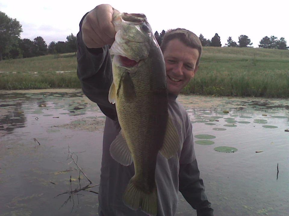 Guy holding a big bass in a pond, trees and grass in background. Supermotors logo at bottom right