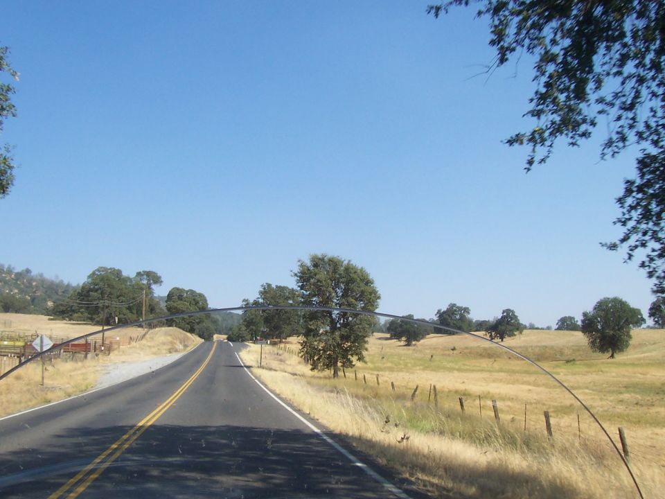 Road scene from inside a vehicle, with dry grass fields and trees under a clear blue sky