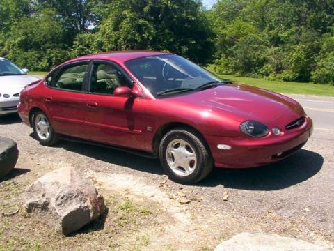 1998 Ford Taurus - A red sedan is parked on a gravel area next to a road with trees in the background.