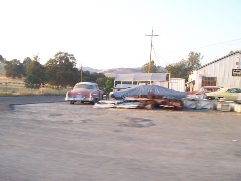 1994 Ford F-150 - A vintage pink car is parked in a lot with other vehicles and covered items near a building.