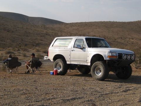 1996 Ford Bronco - A modified white Ford Bronco sits in a desert setting with two people relaxing in camping chairs.