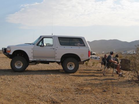 1996 Ford Bronco - A modified white Ford Bronco sits in a desert landscape with people relaxing in chairs nearby.