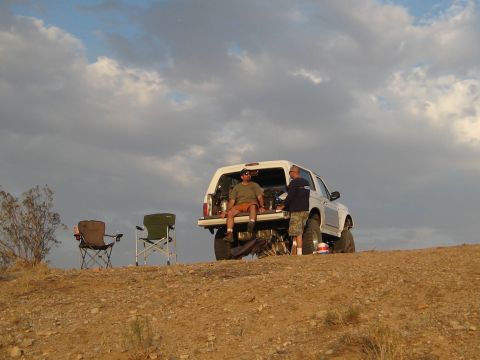 1996 Ford Bronco - Two men are gathered around the open tailgate of a white Ford Bronco parked on a dirt hill. Two camping chairs are set up nearby, suggestin…