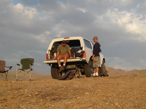 1996 Ford Bronco - Two men are gathered around a white Ford Bronco with its tailgate open, parked in a desert landscape. One man sits on the edge of the tailg…
