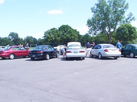 1998 Ford Taurus - A gathering of cars in a parking lot on a sunny day, with several people interacting around the vehicles.