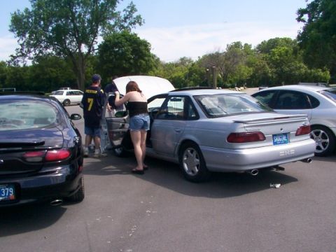 1998 Ford Taurus - A silver sedan is parked next to a black car, with people standing around the open hood of the silver car.