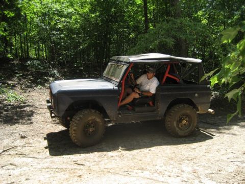 1995 Ford Bronco - Off-road vehicle with roll cage parked on a dirt path surrounded by trees.