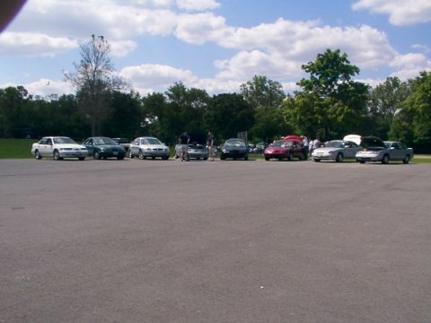 1998 Ford Taurus - A group of sedans are parked in a lot, with some people gathered around a red car.