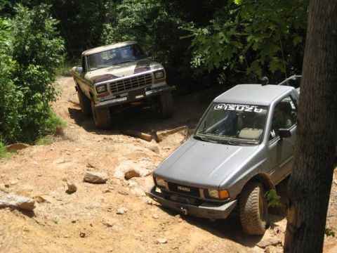 1995 Ford Bronco - Two off-road trucks navigate a challenging dirt trail surrounded by dense foliage.