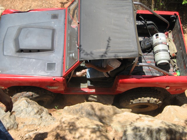 Red off-road vehicle with a gray top and black soft top, driving over rocks. A person is visible in the driver's seat. A propane tank is in the bed