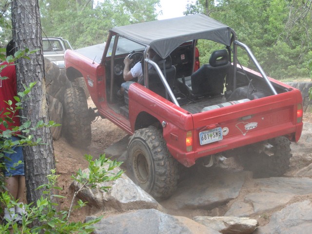 Red SUV with a soft top is going over rocks, another vehicle is behind it