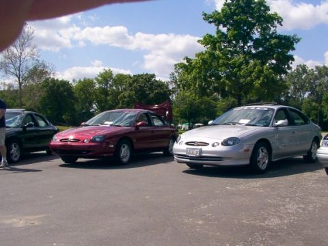 1998 Ford Taurus - Three sedans are parked in a lot on a sunny day with trees in the background.