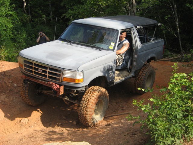 Modified 1995 Ford Bronco with large tires on a dirt trail, driver visible