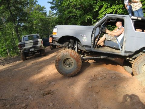 1995 Ford Bronco - Two off-road vehicles navigate a challenging dirt path surrounded by trees.