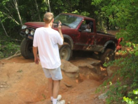 1995 Ford Bronco - A red pickup truck is shown off-roading on a dirt path with large rocks.
