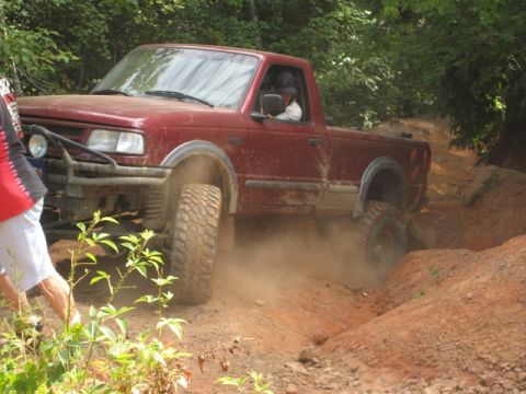 1995 Ford Bronco - A red pickup truck navigates a challenging off-road trail, its tires kicking up a cloud of dust.