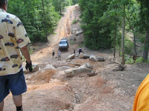 1995 Ford Bronco - Several off-road vehicles are seen on a challenging dirt trail surrounded by trees and large rocks.