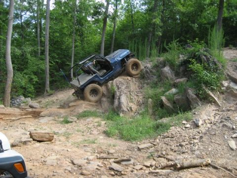 1995 Ford Bronco - A blue off-road jeep navigates a challenging rocky terrain in a wooded area.