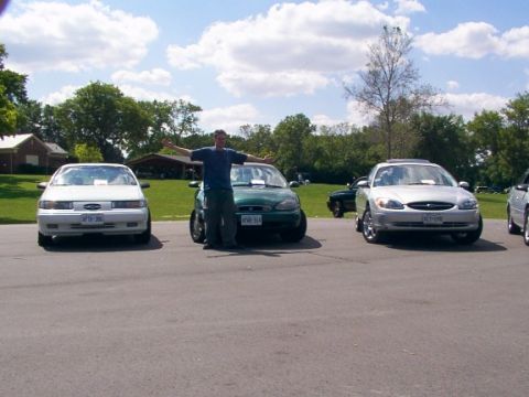 1998 Ford Taurus - A silver 1998 Ford Taurus is parked in front of a gathering of cars, with a person standing between vehicles.