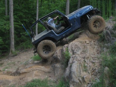 1995 Ford Bronco - A blue off-road jeep navigates a steep, rocky incline in a wooded area.