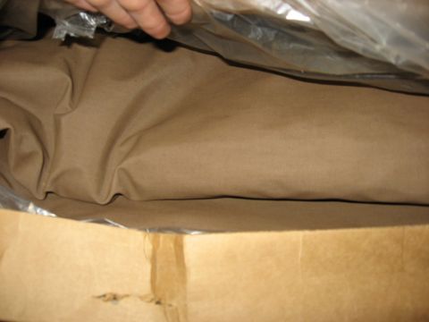 1989 Ford Bronco - Close-up of folded tan canvas-style vinyl fabric still sealed in clear plastic inside an opened cardboard shipping box, with a hand lifting…