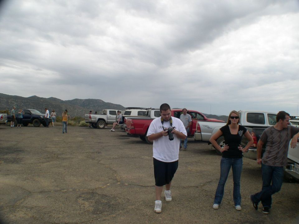 Several pickup trucks parked in a lot with people around. One guy is holding a camera