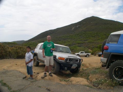 2007 Toyota FJ Cruiser - A group of people and vehicles are gathered on a dirt road with a hilly, vegetated landscape in the background.