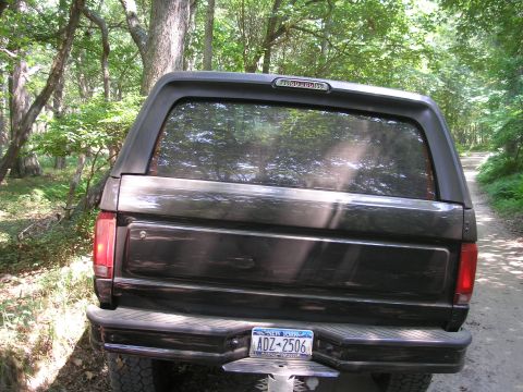 1995 Ford Bronco - The rear of a dark 1995 Ford Bronco with a cap is parked on a dirt road surrounded by trees.