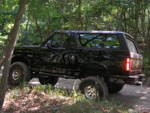 1995 Ford Bronco - A black Ford Bronco is parked on a dirt path surrounded by trees, showing its off-road modifications.
