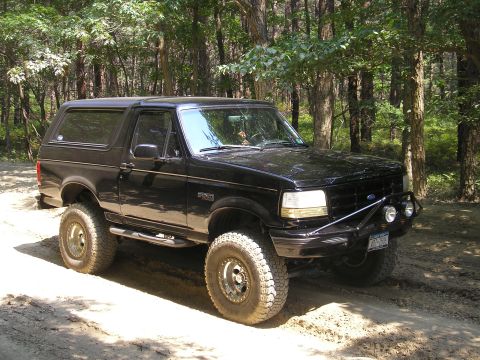 1995 Ford Bronco - This black 1995 Ford Bronco is equipped with off-road tires and a front brush guard, parked on a dirt path surrounded by trees.